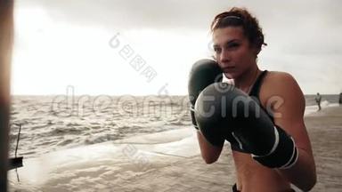 一个强壮的运动女拳击手戴着手套，带着一个袋子在海边晒太阳。 女拳击手